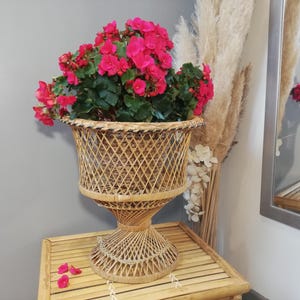 Peut inclure: Un pot en osier tressé avec une base en piédestal, rempli de fleurs roses vibrantes et de feuillage vert. Le pot est posé sur une table en bambou, créant une présentation naturelle et décorative. Le pot mesure environ 61 cm de haut.