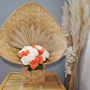 May include: A woven wicker basket with a wide, rounded top, filled with a bouquet of white and orange roses. The basket is sitting on a woven bamboo table. A dried pampas grass arrangement is in a vase to the right.