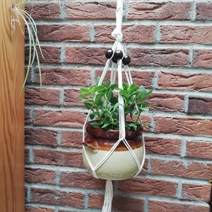 May include: A white macrame plant hanger with black wooden beads hangs from a brick wall. A terracotta pot with a green plant is suspended in the hanger.