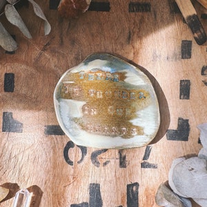 May include: A handcrafted ceramic dish with an irregular form. The dish showcases a mix of earthy colours, including brown, beige, and blue highlights. The text "THERE IS FOUNDER IN OUR HEARTS" is inscribed on the surface. The dish is displayed on a wooden surface.