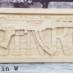 May include: A wooden sign with the name "MCINTROY" carved into it. The sign features a stylized image of a rifle with a wood grain finish. The sign is 23 inches long and 7.25 inches wide and is made of pickled oak.