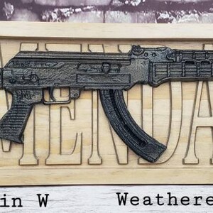 May include: A wooden sign with the word "ARMENDARIZ" carved into it. A black and silver AK-47 style rifle is mounted on the sign. The sign is 23 inches long and 7.25 inches wide. It is made of weathered oak and ebony.