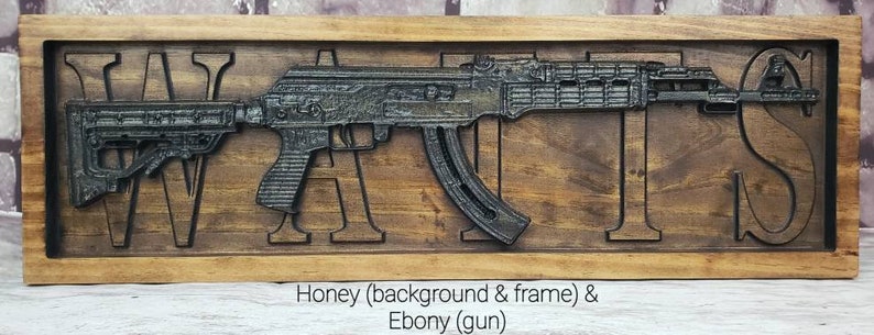 May include: A wooden sign with the word "WANTS" carved into it. A black rifle is carved into the sign, with the barrel extending beyond the letters. The background and frame are honey-colored, while the gun is ebony.