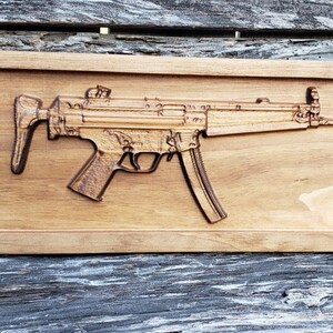 May include: A wooden relief carving of a submachine gun, set within a rectangular frame. The gun is depicted in detail, with its various components clearly visible. The wood has a warm, natural tone.
