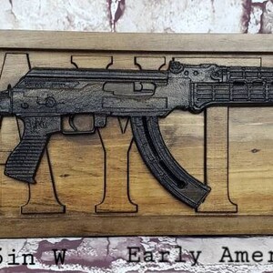 May include: A wooden sign with a black metal cutout of a rifle and the word "DAIN" carved into the wood. The sign is 28 inches long and 9.25 inches wide. It is made of Early American and Ebony wood.