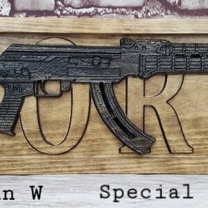 May include: A wooden sign with the name "FLORES" carved into it. A black metal replica of an assault rifle is mounted on the sign. The sign is made of walnut and ebony wood and measures 26 inches long by 7.25 inches wide.