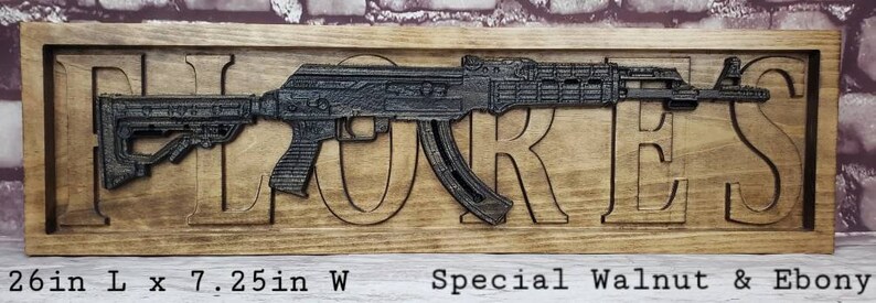 May include: A wooden sign with the name "FLORES" carved into it. A black metal replica of an assault rifle is mounted on the sign. The sign is made of walnut and ebony wood and measures 26 inches long by 7.25 inches wide.