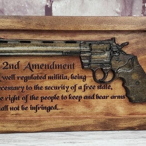 Pode incluir: Uma placa de madeira com uma imagem esculpida de um revólver e o texto "2nd Amendment A well regulated militia, being necessary to the security of a free state, the right of the people to keep and bear arms shall not be infringed..."