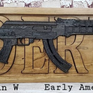 May include: A wooden sign with the word "GORTZ" carved into it. The sign features a black metal replica of an AK-47 rifle. The sign is 28 inches long and 9.25 inches wide. The wood is Early American and Ebony.