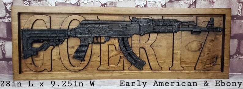 May include: A wooden sign with the word "GORTZ" carved into it. The sign features a black metal replica of an AK-47 rifle. The sign is 28 inches long and 9.25 inches wide. The wood is Early American and Ebony.
