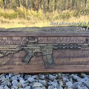 Może przedstawiać: Drewniana tabliczka z rzeźbą karabinu i tekstem "We the People of the United States, in Order to form a more perfect Union..." Tabliczka jest wykończona w kolorze czerwonego mahoniu i hebanu. Ma 56 cm długości i 18,4 cm szerokości.