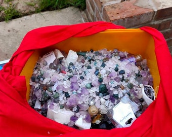 Puede incluir: Un contenedor de plástico amarillo lleno de una variedad de cristales y piedras, incluyendo amatista, cuarzo transparente y piedras verdes. El contenedor está forrado con una camiseta roja.
