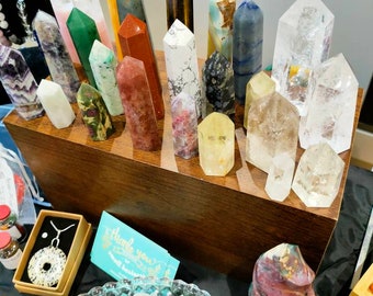 Puede incluir: Una colección de puntas y varitas de cristal de varios colores y formas, incluyendo cuarzo transparente, amatista, aventurina verde, ojo de tigre y cuarzo rosa. Los cristales están dispuestos en un estante de madera y un cuenco de cristales más pequeños está en primer plano.