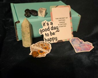 Puede incluir: Una caja turquesa con una tarjeta blanca y rosa que dice "El secreto para salir adelante es empezar." Hay una pegatina que dice "Es un buen día para ser feliz" con girasoles y una piedra marrón. Hay un cristal verde, un cristal blanco y un cristal morado en la caja.