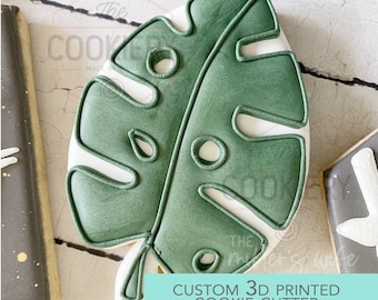 May include: A green 3D printed cookie cutter shaped like a monstera leaf. The cutter has a white outline and several holes cut out of the leaf. The text "CUSTOM 3D PRINTED COOKIE CUTTER" is printed on a blue background below the cutter.