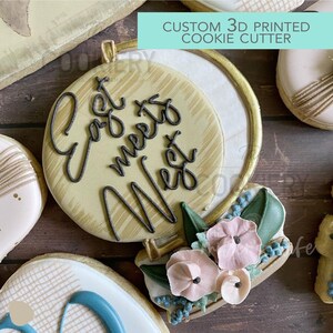 May include: A 3D printed cookie cutter shaped like a globe with the words "Go West" written on it. The globe is decorated with a floral arrangement of pink and blue flowers and green leaves.