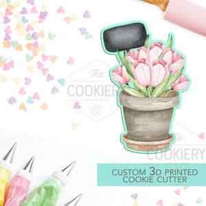 May include: A 3D printed cookie cutter in the shape of a potted plant with pink tulips and a blank sign. The cutter is made by The Cookiery, Made by Mama.