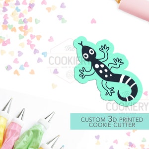 May include: A turquoise blue 3D printed cookie cutter shaped like a black and white lizard with a striped tail. The cutter is on a white background with colourful heart sprinkles. The text "Custom 3D Printed Cookie Cutter" is below the cutter.
