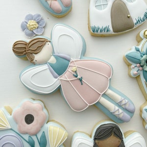 May include: A blue and pink fairy cookie with white wings, brown hair, and a flower on her dress. The cookie is decorated with white icing and has a light blue background.