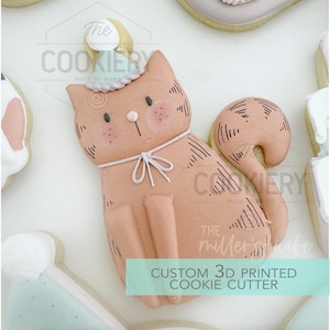 May include: A 3D printed cookie cutter in the shape of a cat wearing a party hat. The cutter is made of white plastic and has a detailed design. The text "The Cookiery Made by Mama" is visible on the cutter.