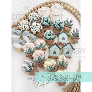 May include: A collection of decorated cookies in various shapes, including birds, flowers, and birdhouses. The cookies are arranged on a white surface with wooden beads and a rope. The text "CUSTOM 3D PRINTED COOKIE CUTTER" is visible.
