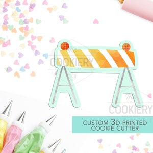 May include: A 3D printed cookie cutter shaped like a construction barrier, featuring orange and white stripes. The text "CUSTOM 3D PRINTED COOKIE CUTTER" is below the cutter. Sprinkles and icing bags are in the background.