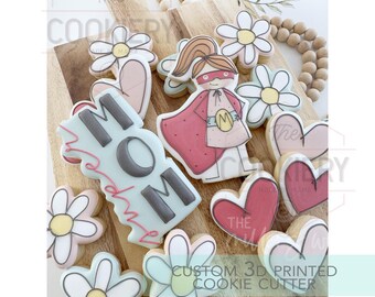 May include: A set of 3D printed cookie cutters featuring a superhero mom, a heart, and a daisy. The cutters are made of white plastic and are designed to cut out cookies in the shape of a superhero mom, a heart, and a daisy. The cutters are shown on a wooden surface with other decorated cookies.