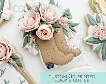 May include: A custom 3D printed cookie cutter in the shape of cowboy boots with a floral arrangement. The boots are tan, and the flowers are pink and white. The text "CUSTOM 3D PRINTED COOKIE CUTTER" is visible.