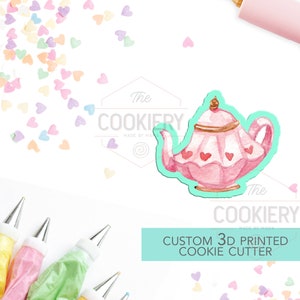 Puede incluir: Un cortador de galletas de tetera rosa y blanca con detalles de corazón. El cortador está impreso en 3D y es parte de un conjunto de cortadores de galletas personalizados. La imagen también muestra mangas pasteleras y chispas.