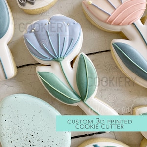 Könnte beinhalten: Ein 3D-gedruckter Ausstecher in Form einer blauen Blume mit grünen Blättern. Der Text "CUSTOM 3D PRINTED COOKIE CUTTER" ist auf blauem Hintergrund gedruckt.