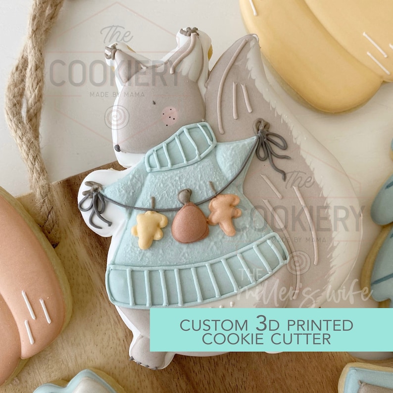 May include: A 3D printed cookie cutter in the shape of a squirrel wearing a blue sweater and holding a garland of fall leaves. The text "CUSTOM 3D PRINTED COOKIE CUTTER" is printed below the squirrel.