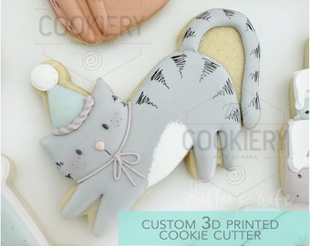 May include: A grey cat wearing a blue and white party hat with a white pom-pom on top. The cat is wearing a pink bow around its neck. The cat is a 3D printed cookie cutter.
