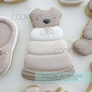 May include: A 3D printed cookie cutter shaped like a stack of rings with a bear head on top. The cookie cutter is made of white plastic and has a light brown colour. The text "CUSTOM 3D PRINTED COOKIE CUTTER" is printed on the bottom of the cookie cutter.