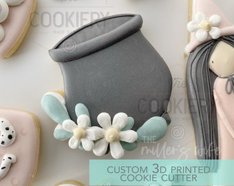 Può includere: Un tagliapasta grigio stampato in 3D a forma di calderone con fiori bianchi e foglie verdi attorno al bordo. Il testo "CUSTOM 3D PRINTED COOKIE CUTTER" è nella parte inferiore dell'immagine.