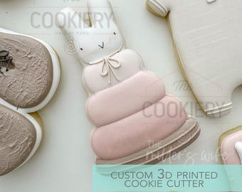 May include: A 3D printed cookie cutter in the shape of a pink and white stacking toy with a bunny rabbit on top. The cutter is labeled "Custom 3D Printed Cookie Cutter".