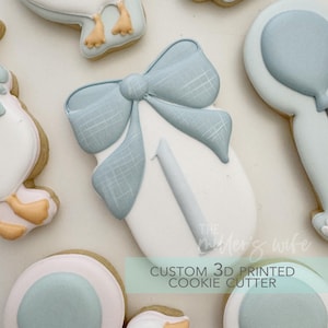 Puede incluir: Primer plano de varias galletas con temática de bebé. Una galleta tiene forma de bebé envuelto en una manta con un lazo azul. Otras galletas incluyen un sonajero y una cigüeña. Las galletas están decoradas con glaseado blanco, azul claro y beige.