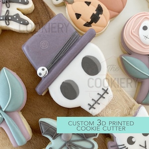 May include: A 3D printed cookie cutter shaped like a skull wearing a purple hat. The cookie cutter is on a wooden surface with other decorated cookies. The text "CUSTOM 3D PRINTED COOKIE CUTTER" is on the image.