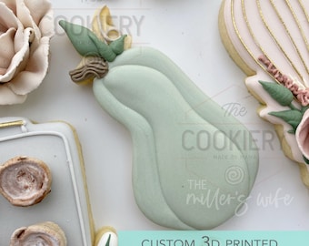 May include: A 3D printed cookie cutter in the shape of a green pear with a brown stem and leaves. The cutter is on a white surface with the text "CUSTOM 3D PRINTED COOKIE CUTTER" below it.