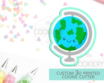 May include: A custom 3D printed cookie cutter in the shape of a smiling globe with blue oceans and green continents. The cutter is outlined in a light green color. Sprinkles and frosting bags are in the background.