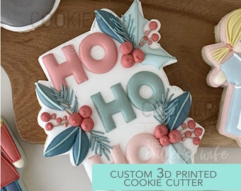 Puede incluir: Un cortador de galletas blanco con las palabras "HO HO" en glaseado rosa y azul. El cortador de galletas está decorado con hojas verdes y bayas rojas. El texto "CUSTOM 3D PRINTED COOKIE CUTTER" está escrito debajo del cortador de galletas.