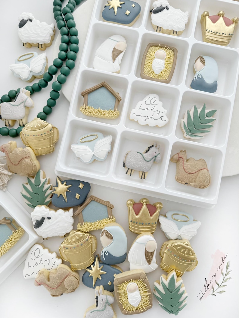K&ouml;nnte beinhalten: Ein wei&szlig;es Tablett gef&uuml;llt mit verzierten Zuckerkeksen mit einem Krippen-Motiv. Die Kekse sind in verschiedenen Farben verziert, darunter Blau, Gold und Wei&szlig;. Die Kekse zeigen Bilder von einem Stern, einer Krippe, einem Kamel, einem Esel, einem Hirten, einem Schaf und einem Engel. Das Tablett ist von einer Vielzahl anderer Kekse umgeben, darunter ein Stern, eine Krippe, ein Kamel, ein Esel, ein Hirten, ein Schaf und ein Engel.