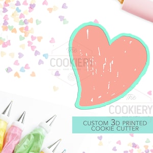 May include: A custom 3D printed cookie cutter in the shape of a heart. The cutter is light pink with a teal outline. The cutter is surrounded by colorful heart-shaped sprinkles and a rolling pin. The text "The Cookiery" is visible in the image.