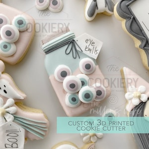 May include: A 3D printed cookie cutter shaped like a mason jar filled with eyeballs. The cookie cutter is pink and blue with a tag that says "eye balls". The cookie cutter is part of a set of Halloween cookie cutters.