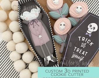 Può includere: Un set di stampini per biscotti stampati in 3D che presenta un vampiro in una bara, un teschio in una bara e tre fantastici disegni di fiori. Gli stampini per biscotti sono neri e bianchi con accenti rosa e verdi. Il testo "Trick or Treat Yourself" è stampato sullo stampino per biscotti a forma di bara.