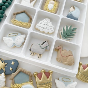 K&ouml;nnte beinhalten: Ein wei&szlig;es Tablett gef&uuml;llt mit weihnachtlichen Zuckerpl&auml;tzchen. Die Pl&auml;tzchen sind mit verschiedenen Bildern verziert, darunter ein Stern, eine Krippe, ein Kamel, ein Schaf, eine Krone und ein Engel. Die Pl&auml;tzchen sind in einem Raster angeordnet. Das Tablett befindet sich auf einer wei&szlig;en Oberfl&auml;che.