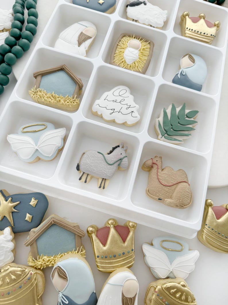 K&ouml;nnte beinhalten: Ein wei&szlig;es Tablett gef&uuml;llt mit weihnachtlichen Zuckerpl&auml;tzchen. Die Pl&auml;tzchen sind mit verschiedenen Bildern verziert, darunter ein Stern, eine Krippe, ein Kamel, ein Schaf, eine Krone und ein Engel. Die Pl&auml;tzchen sind in einem Raster angeordnet. Das Tablett befindet sich auf einer wei&szlig;en Oberfl&auml;che.