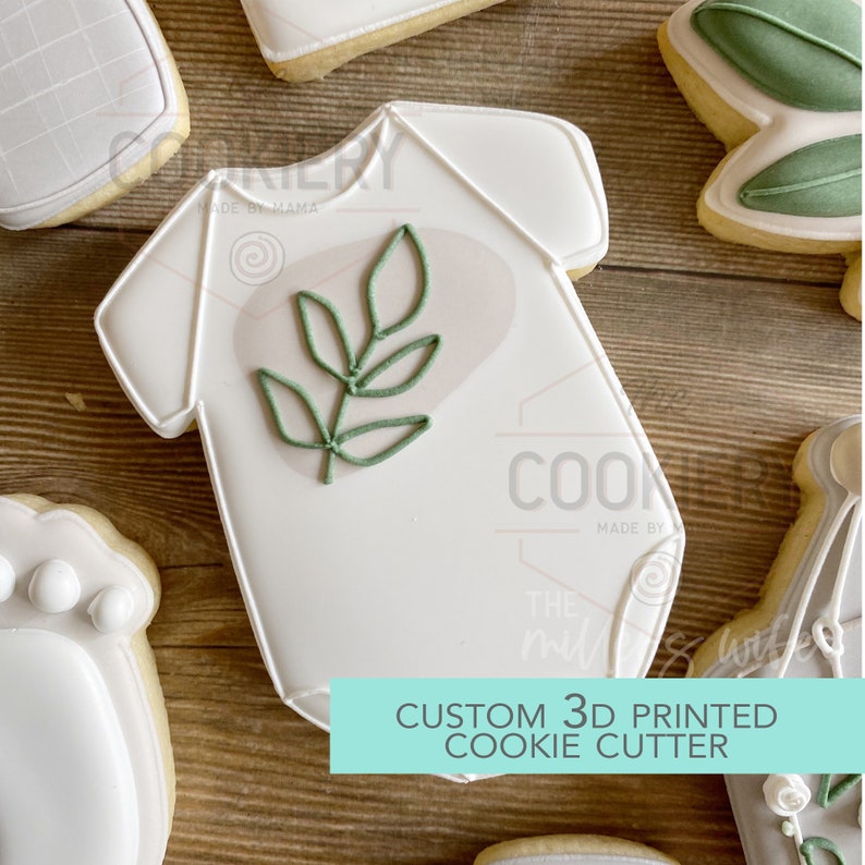 May include: A white cookie cutter in the shape of a baby onesie with a green leaf design. The cookie cutter is 3D printed and says "Custom 3D Printed Cookie Cutter" on the bottom.