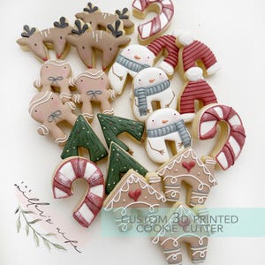 May include: A selection of festive Christmas cookies, featuring reindeer, gingerbread figures, snowmen, Christmas trees, candy canes, and houses. The cookies are decorated with white, red, green, and brown icing. The text "CUSTOM 3D PRINTED COOKIE CUTTER" is visible.