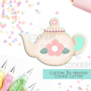 Peut inclure: Un emporte-pièce à biscuits personnalisé imprimé en 3D en forme de théière avec une fleur rose et des feuilles vertes. L'emporte-pièce est fabriqué par The Cookiery, Made by Mama. L'image montre également des poches à douille et des vermicelles.