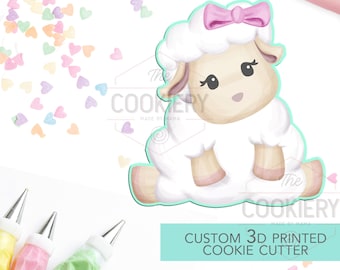 May include: A white 3D printed cookie cutter shaped like a sitting lamb with a pink bow. The cutter is on a white background with colourful heart sprinkles and piping bags.